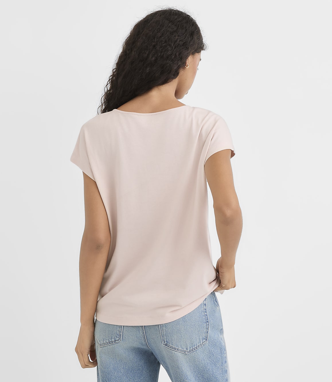 A woman in a light pink t-shirt and blue jeans stands against a plain white background.