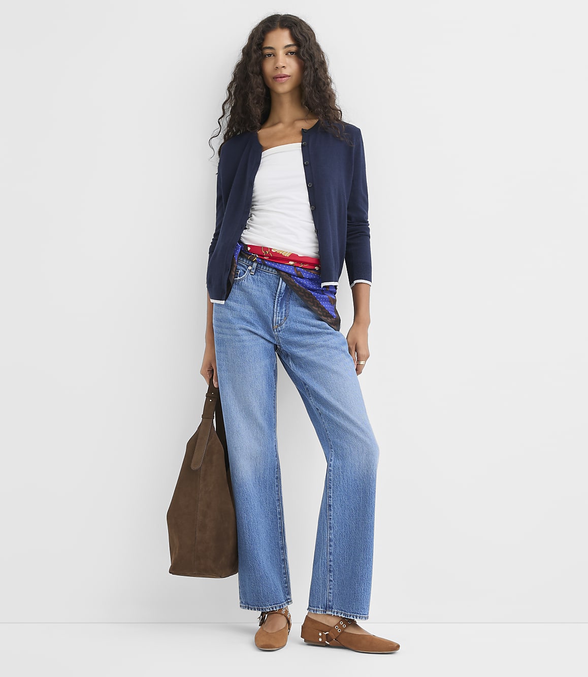 A woman in a white top, navy cardigan, and wide-leg jeans stands against a plain white background, holding a brown suede bag.