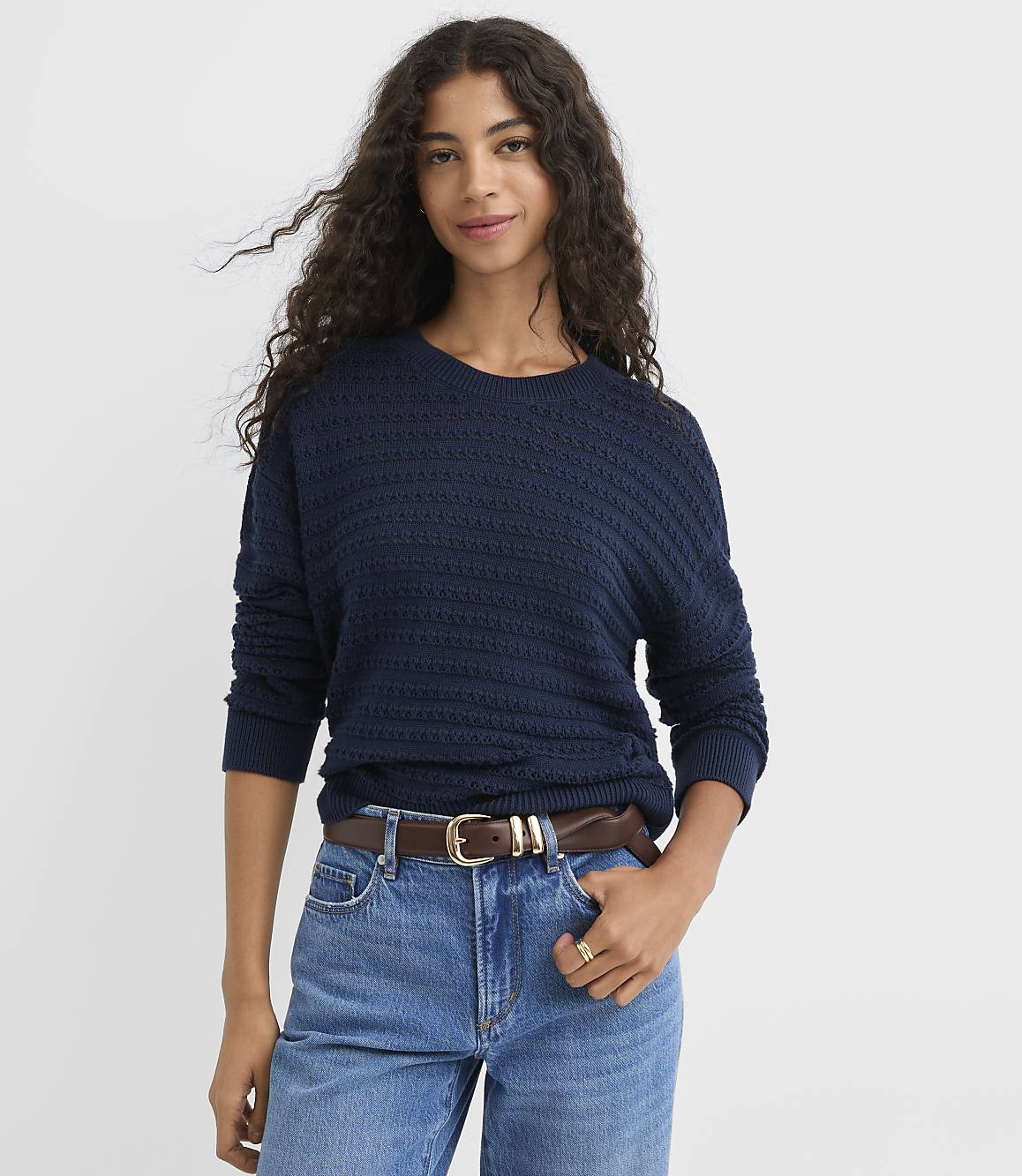 A woman in a navy blue sweater and jeans stands against a plain white background.