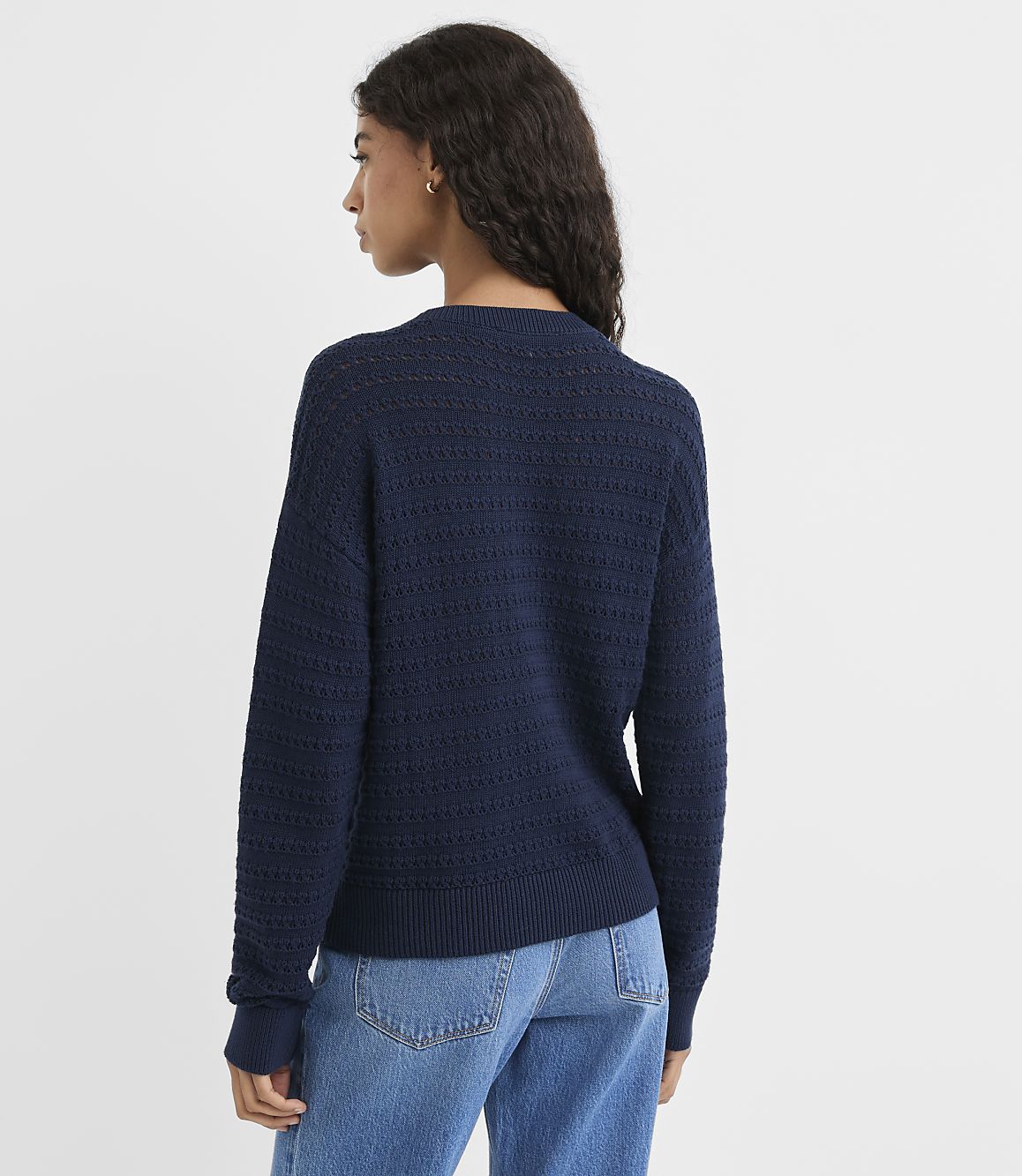 A woman in a navy blue textured sweater and jeans stands against a plain white background.
