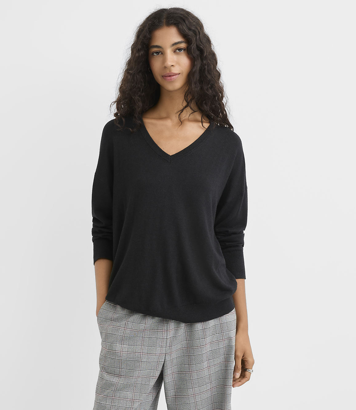 A woman in a black v-neck sweater and plaid pants stands against a plain white background.