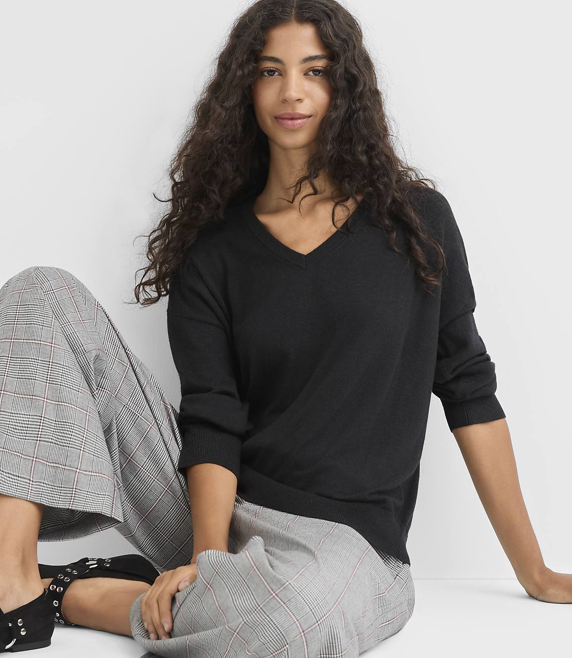 A woman with long, dark, curly hair sits in the foreground wearing a black v-neck sweater and plaid pants, with a plain white background behind her.