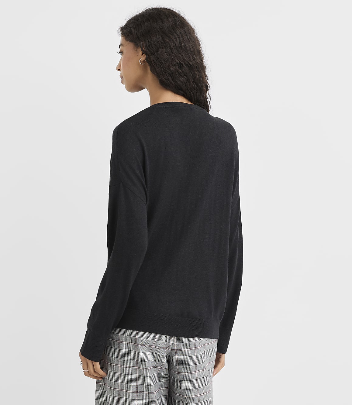 A woman in a black sweater and plaid pants stands against a plain white background.
