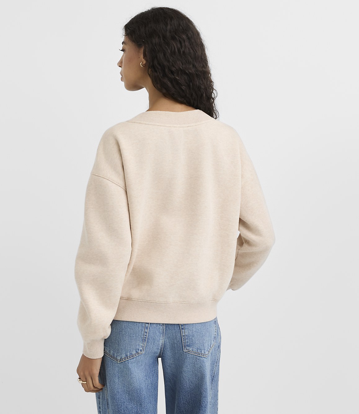 A woman in a cream sweatshirt and blue jeans stands against a plain, light background.