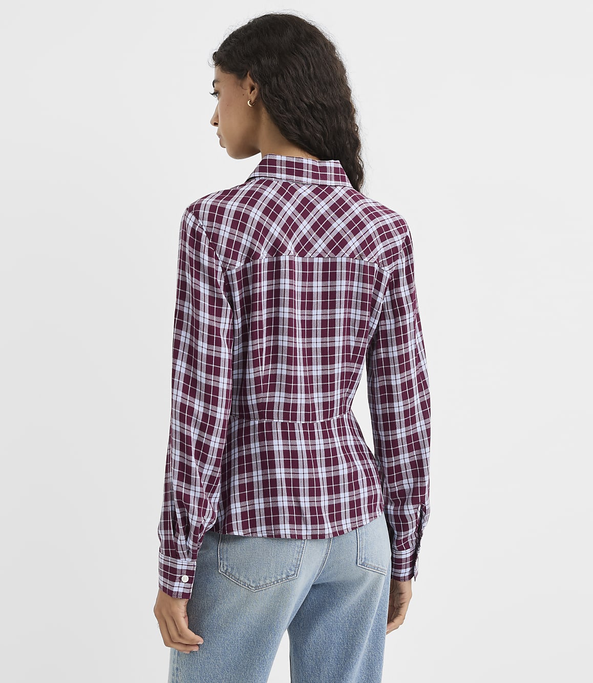 A woman in a maroon and white plaid shirt and blue jeans stands against a plain white background.