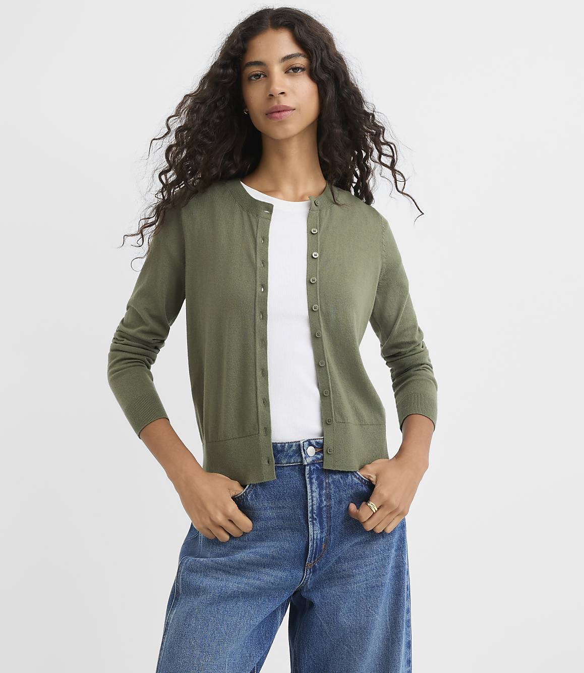 A woman in a green cardigan and blue jeans stands against a plain white background.