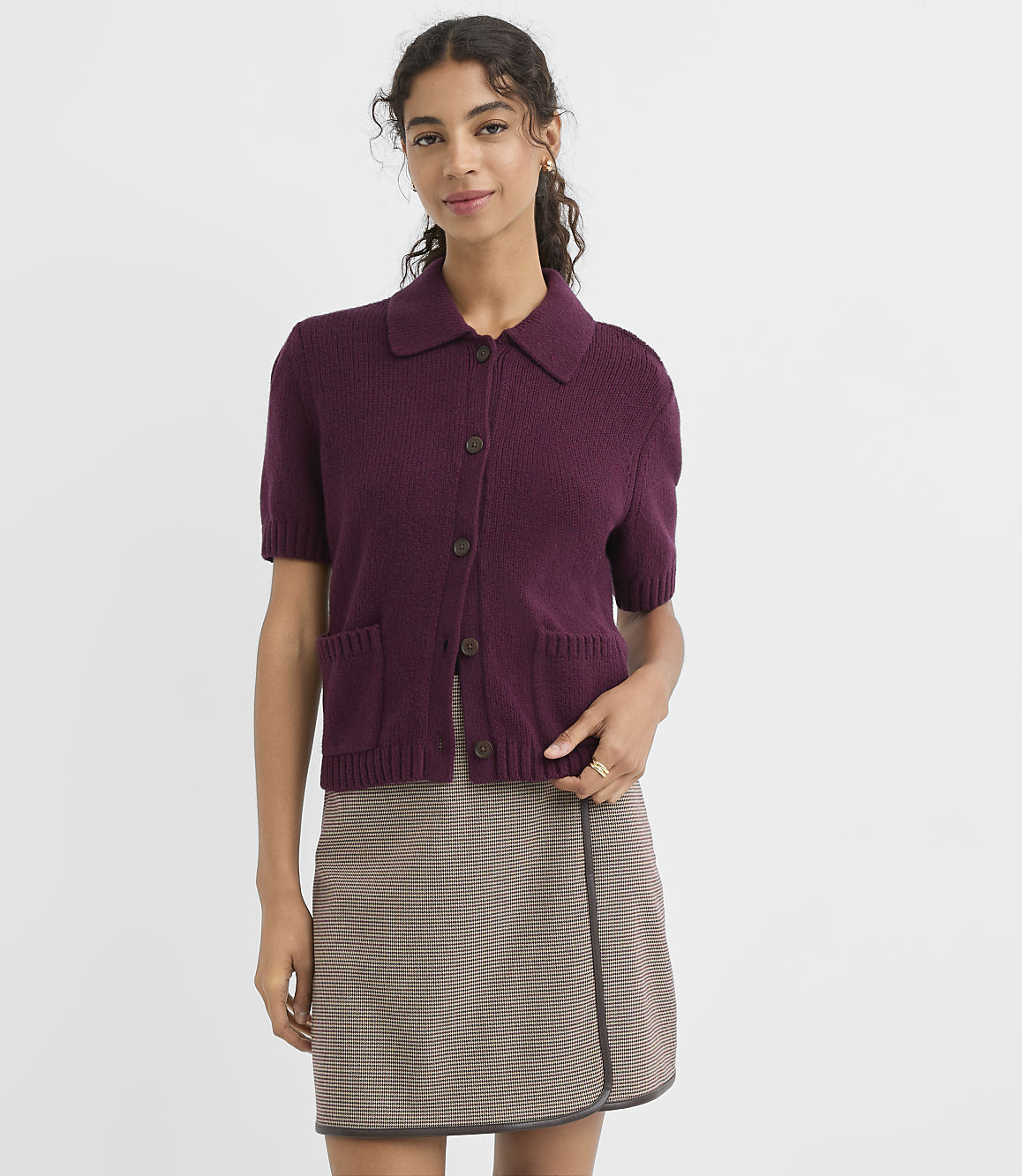 A woman in a burgundy collared cardigan and a plaid skirt stands against a plain white background.