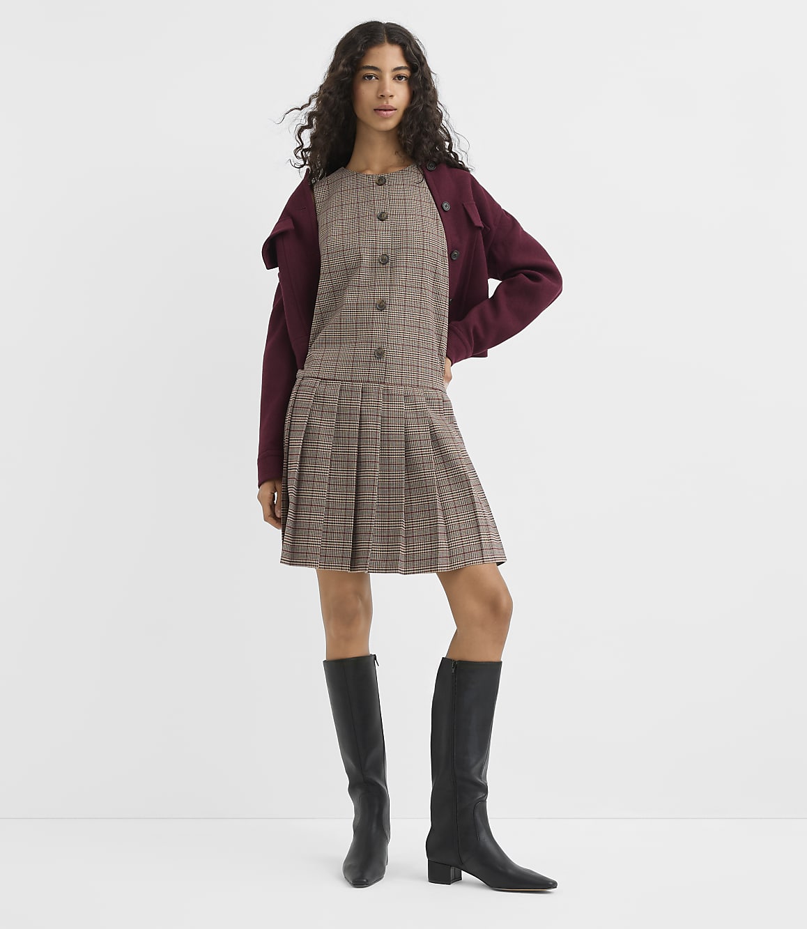 A young woman in a plaid dress and black boots stands against a plain white background.