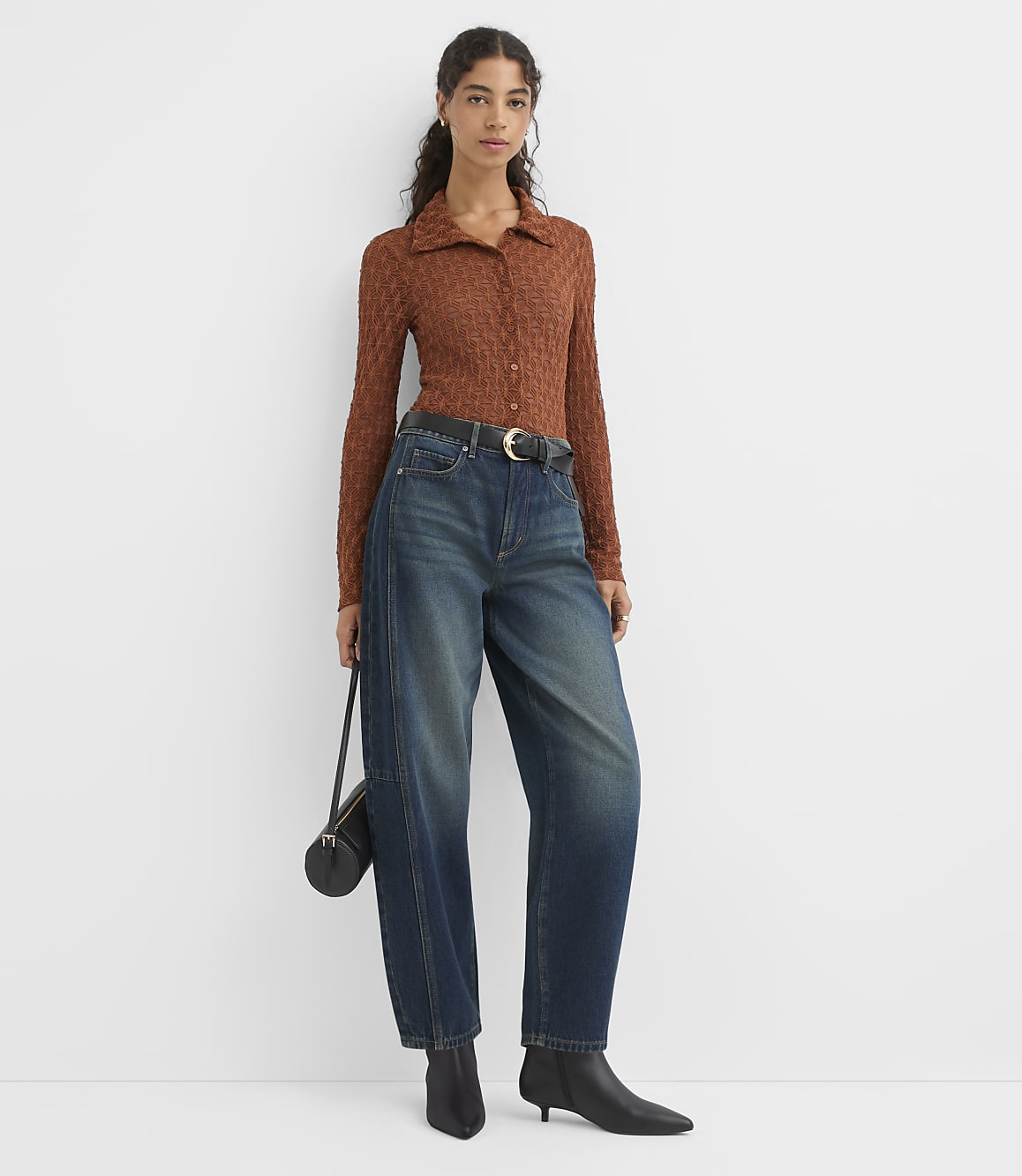 A woman in a brown textured collared shirt, dark wash wide-leg jeans, and black ankle boots stands against a plain white background, holding a black purse.