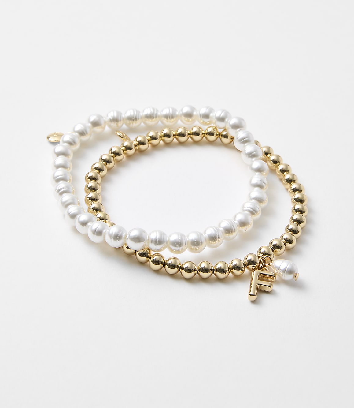 Two bracelets, one with pearls and the other with gold beads and an "F" charm, rest on a plain white background.