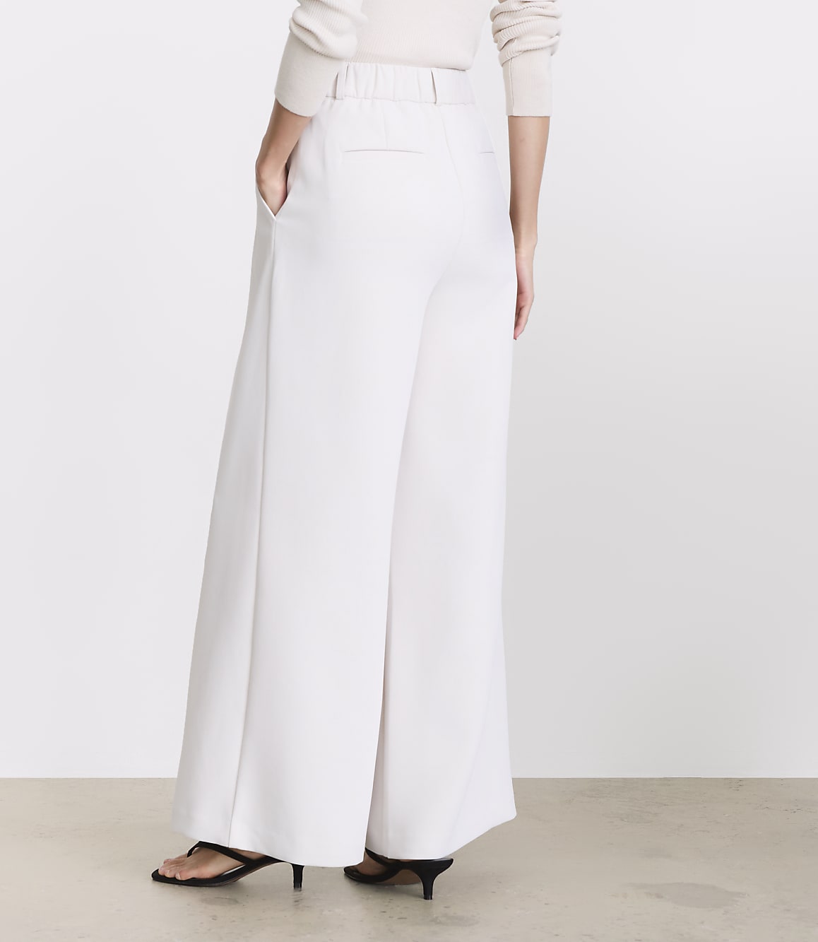 A person wearing white wide-leg pants and black heels stands against a plain, light-colored background.