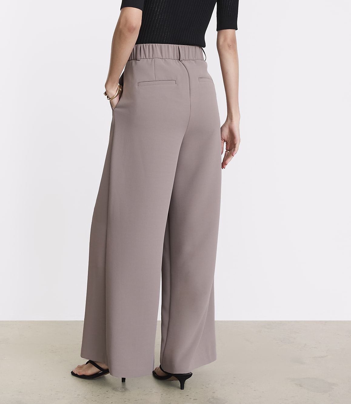 A person wearing wide-leg taupe pants and a black top stands with their back to the camera against a plain white background.