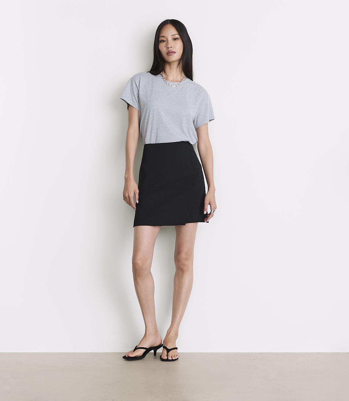A woman in a grey t-shirt and black skirt stands against a plain white background.