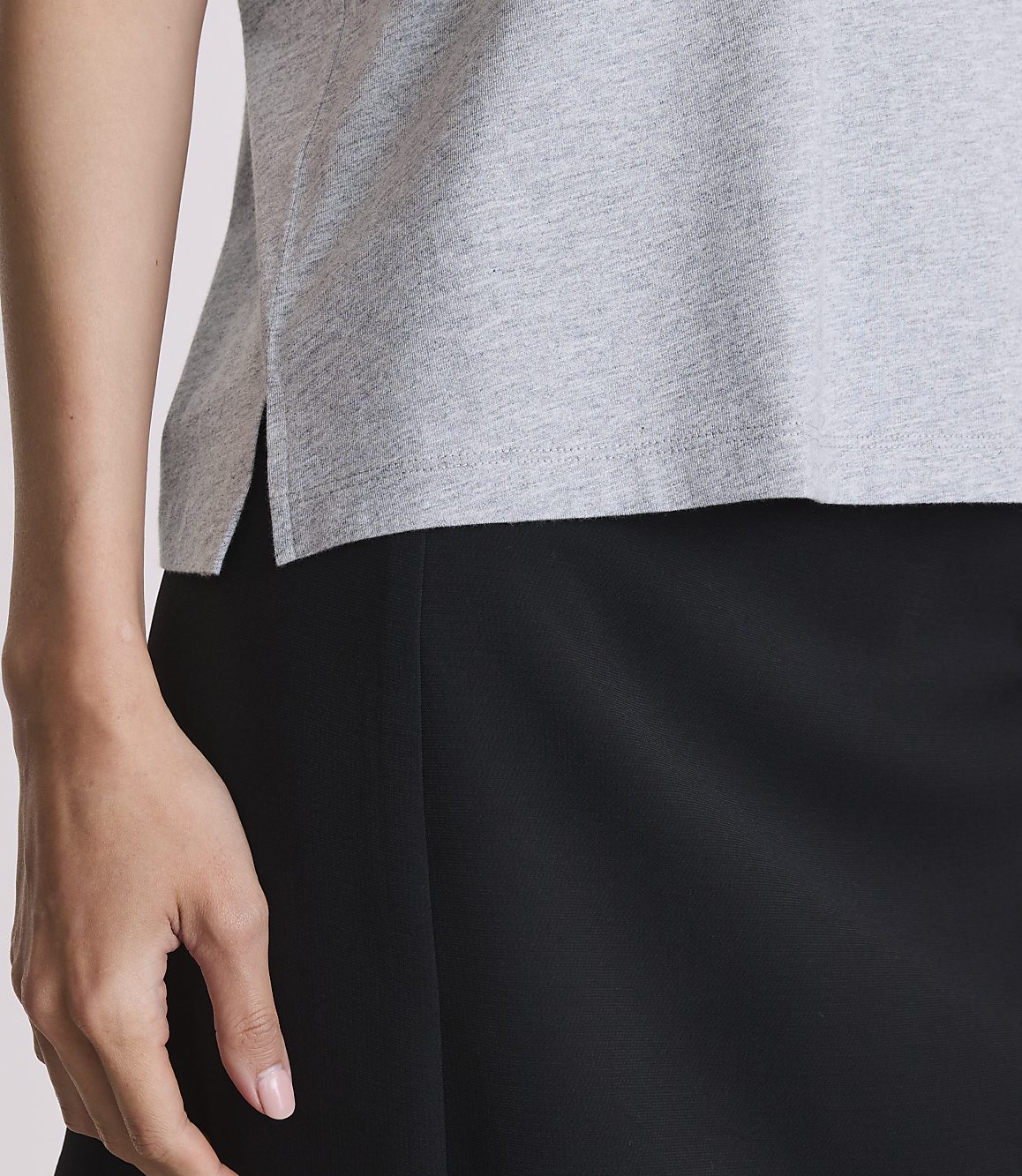 A woman's arm and shoulder in a grey t-shirt are in the foreground, with a black skirt below and a plain background.