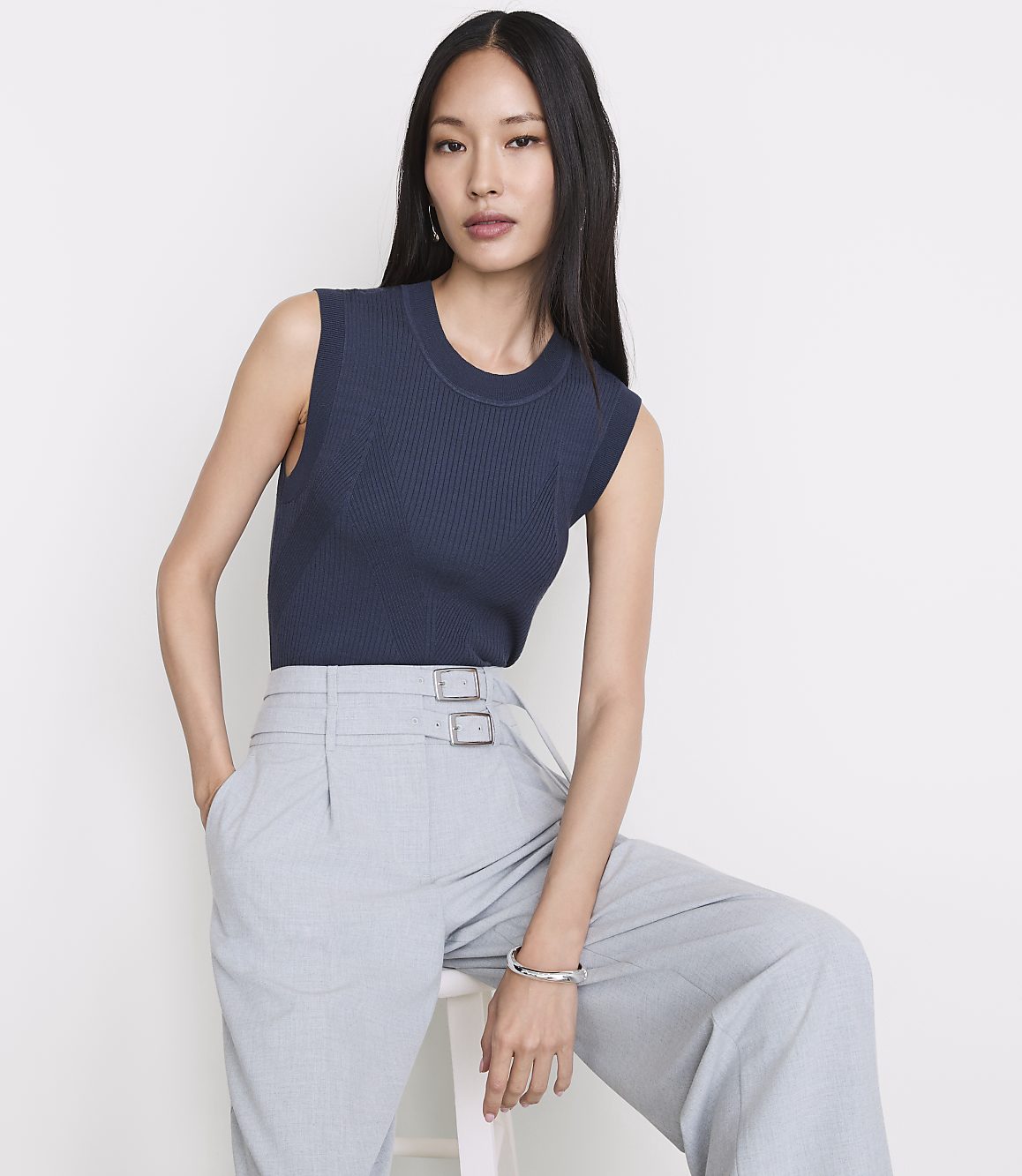 A woman in a navy ribbed tank top and light gray pleated pants sits against a plain white background.