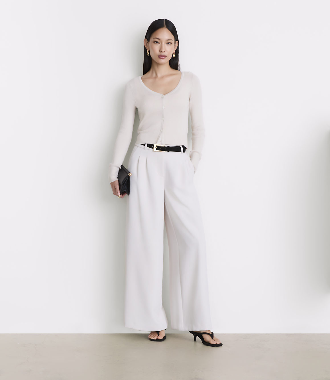 A woman in a cream cardigan and white wide-leg pants stands against a plain white background.