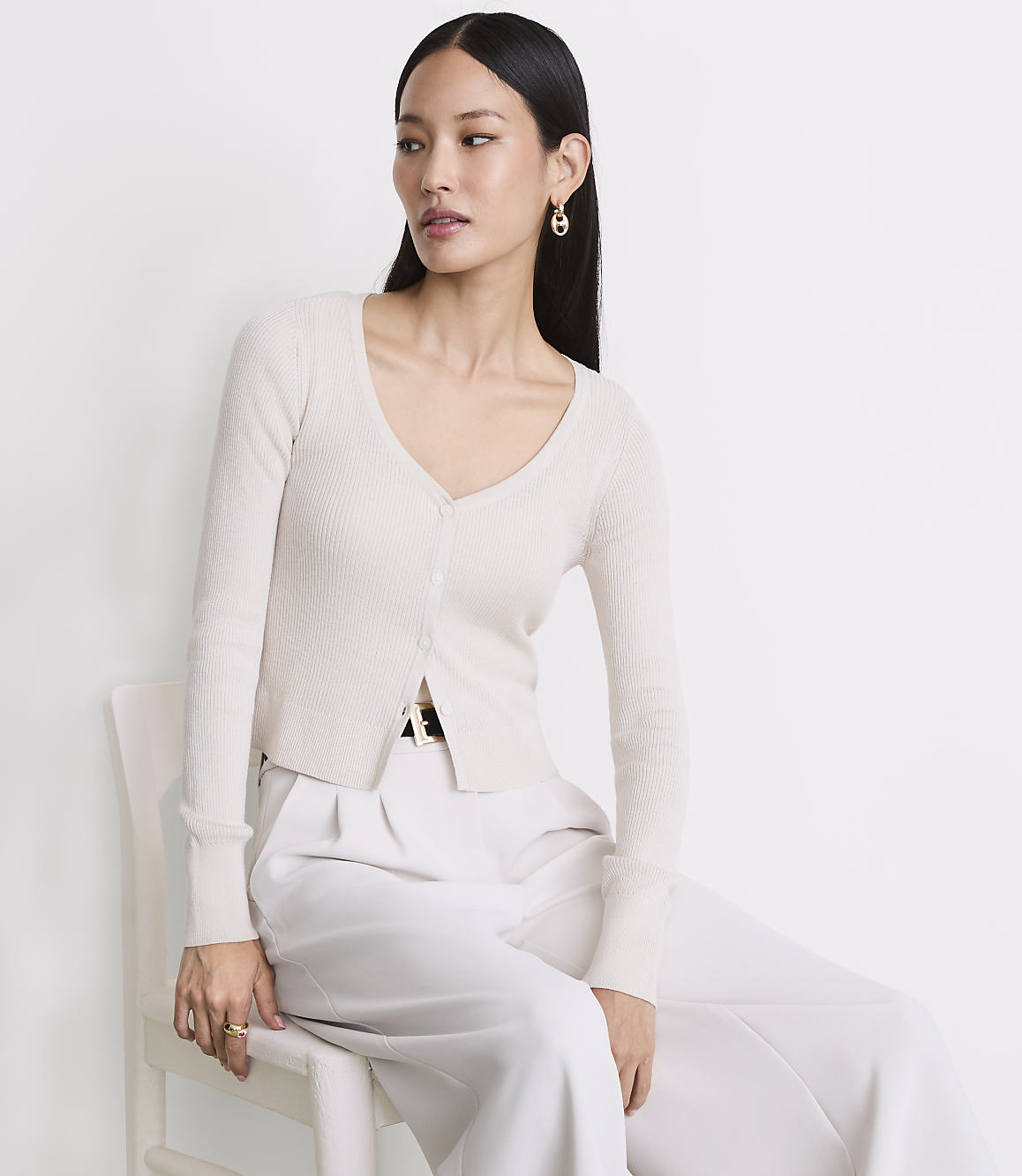 A woman in a cream cardigan and white pants sits on a white chair against a plain white background.