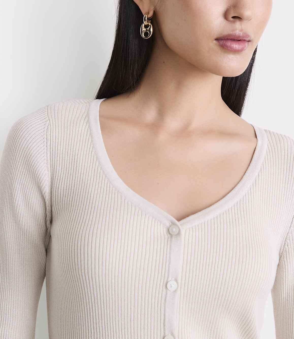 A woman with dark hair and gold earrings wears a cream-colored ribbed cardigan against a plain white background.