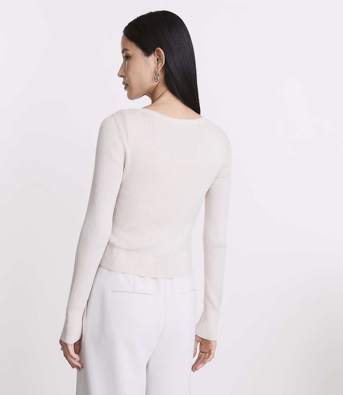 A woman in a cream-colored ribbed sweater and white pants stands against a plain white background.