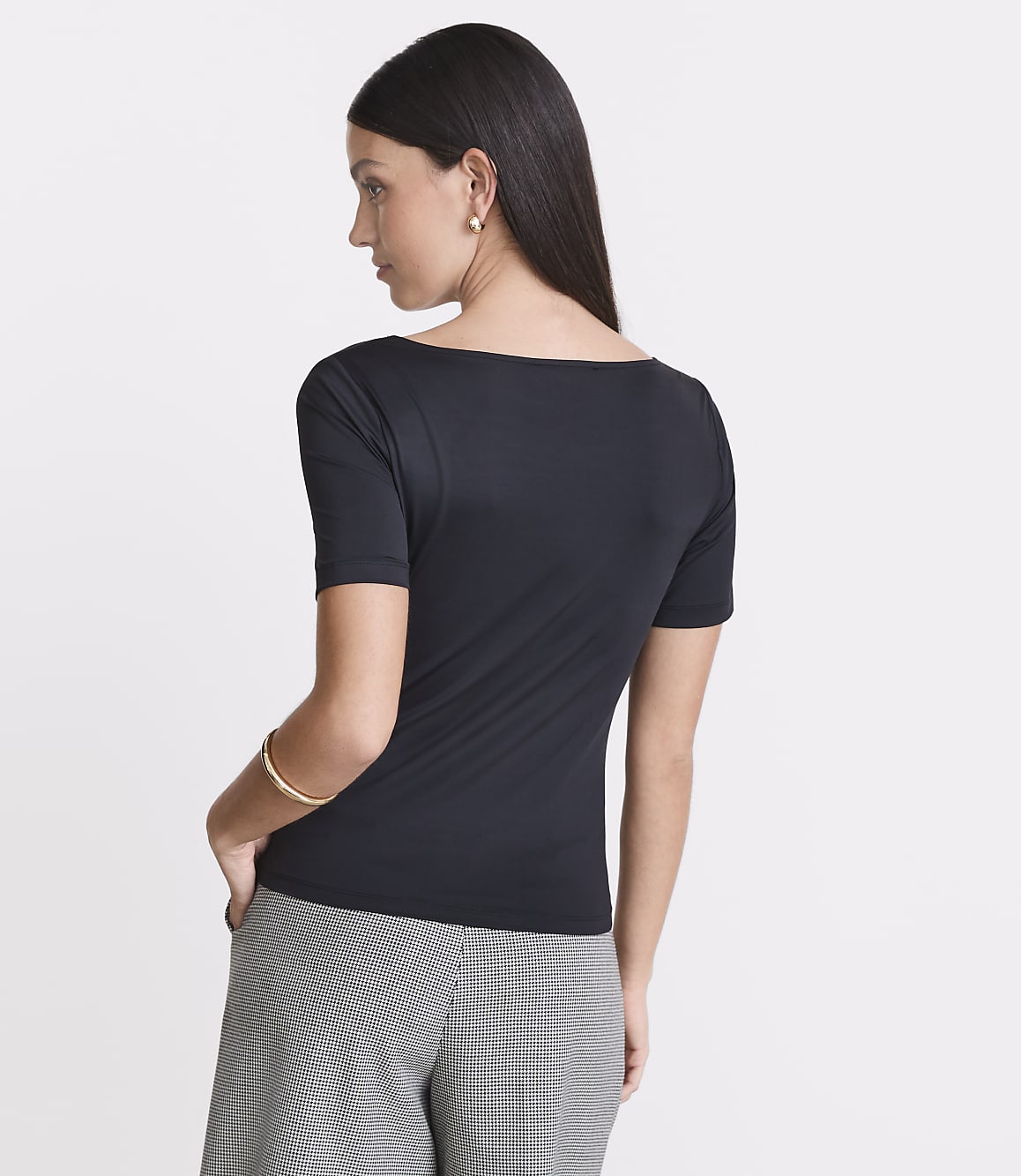 A woman in a black boatneck shirt and checkered pants stands against a plain white background.
