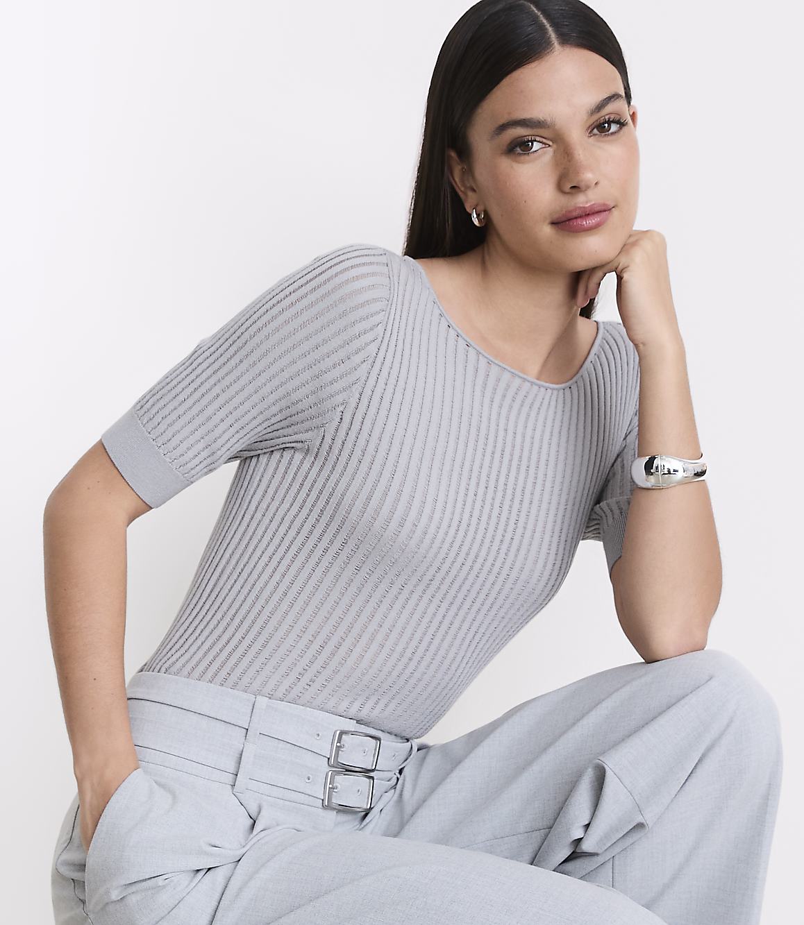 A woman in a grey ribbed top and light blue pants poses against a plain white background.