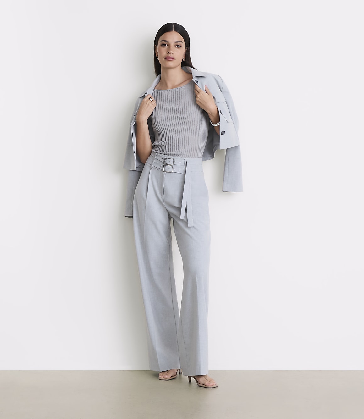 A woman in a grey ribbed top, light blue belted trousers, and a light blue jacket draped over her shoulders stands against a plain white background.