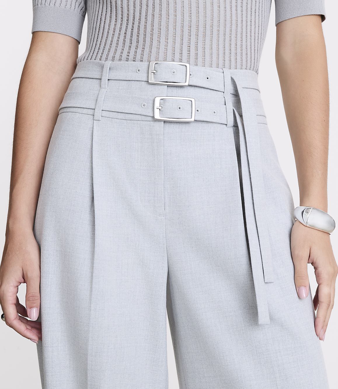 A person in a grey ribbed top and light grey pleated pants with a double buckle belt stands against a plain white background.