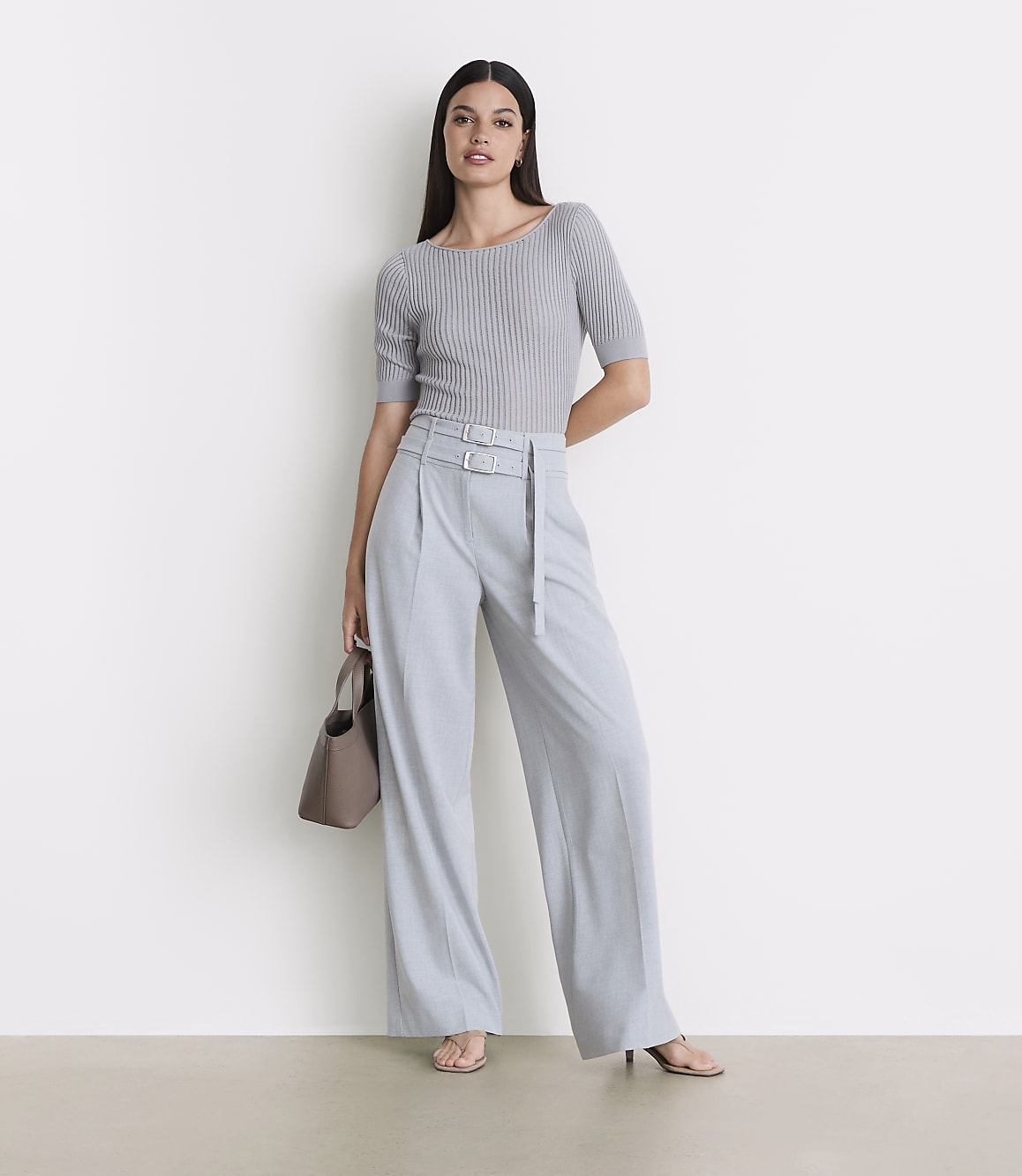 A woman in a grey ribbed top and light blue wide-leg pants stands against a plain white background, holding a taupe handbag.