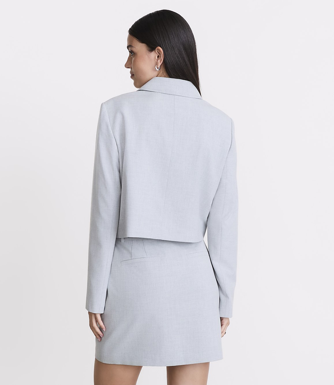 A woman in a light gray cropped blazer and skirt stands against a plain white background.
