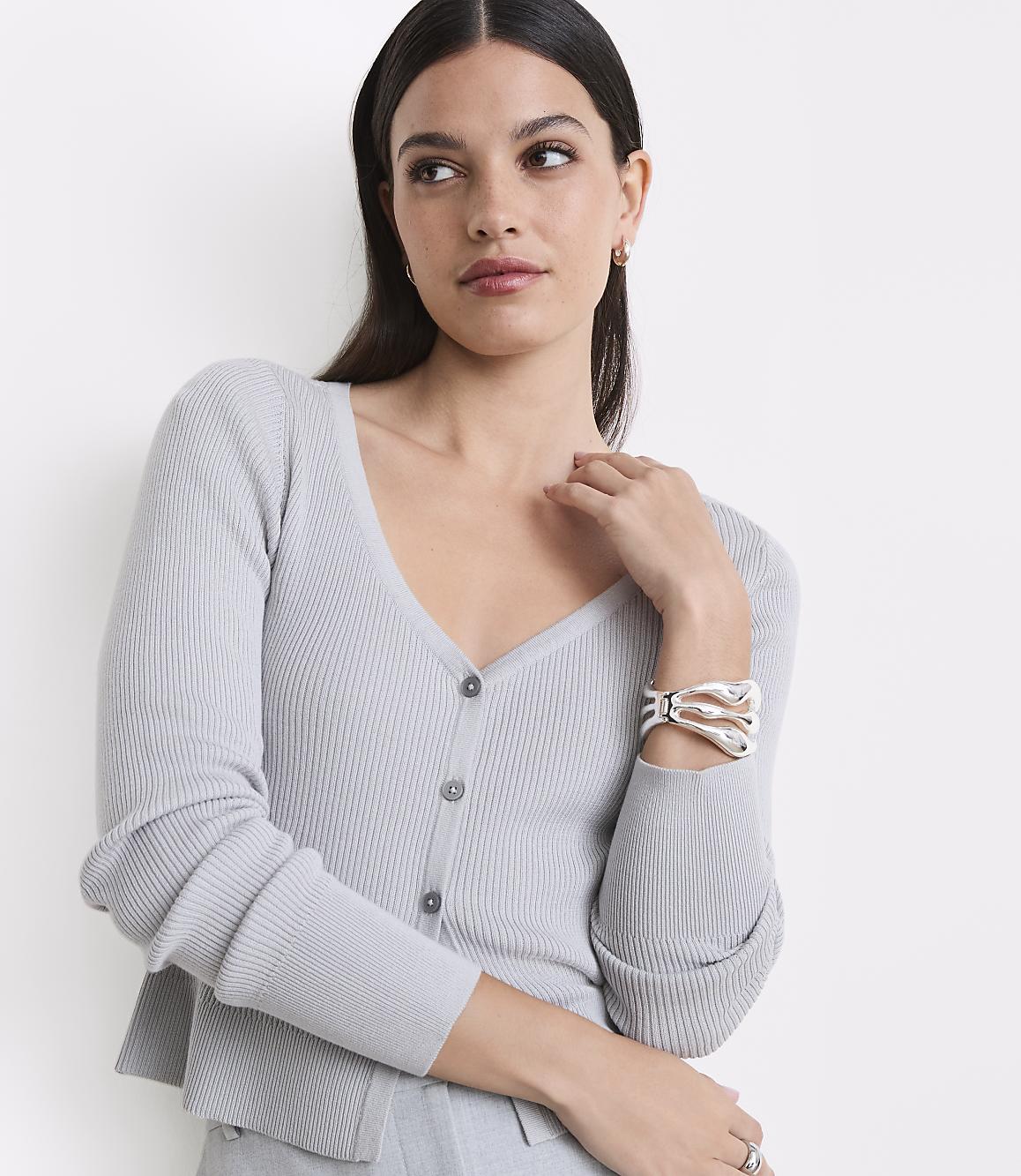 A woman in a light gray ribbed cardigan and bracelet poses against a plain white background.