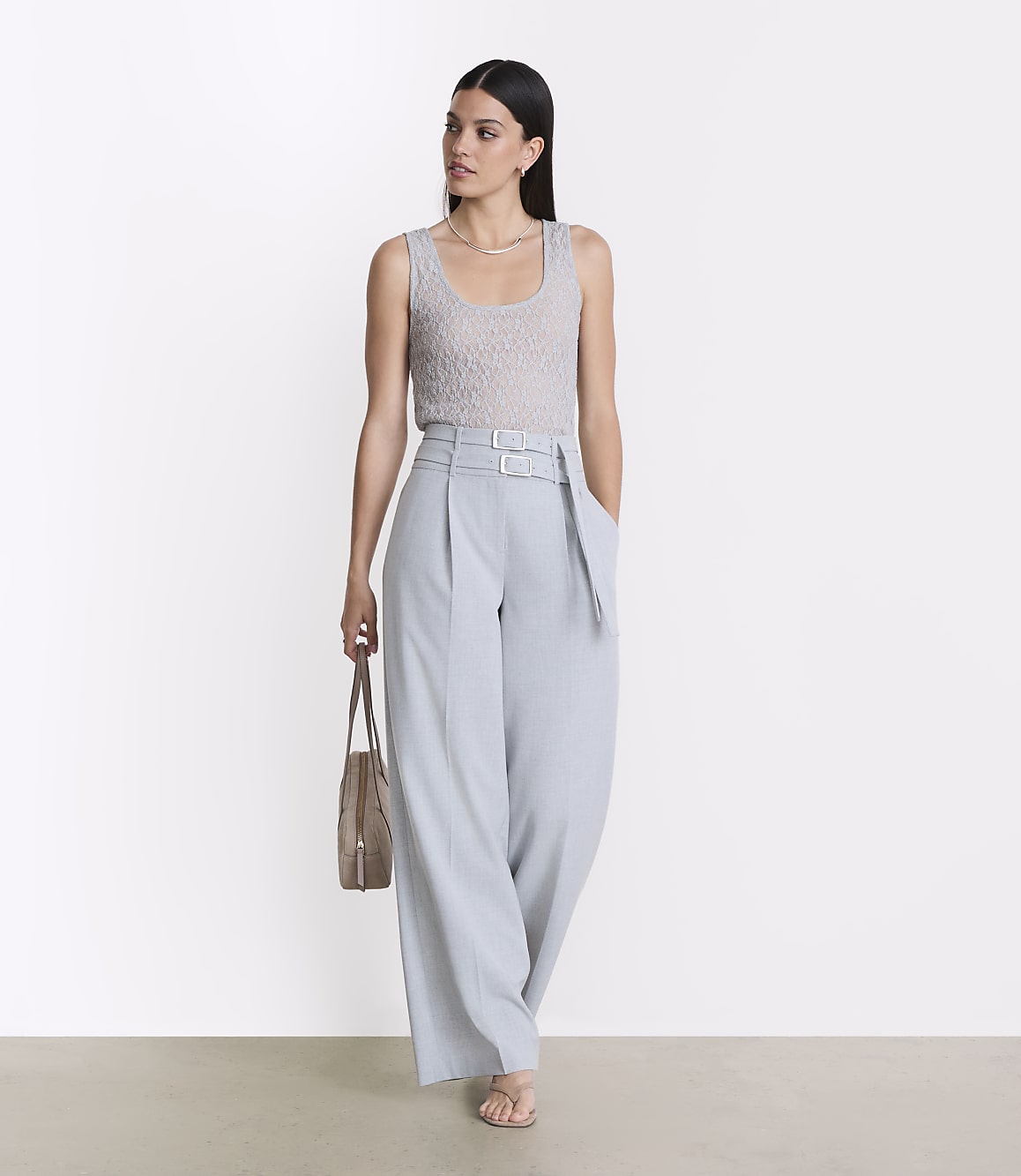 A woman in a light gray tank top and pale blue wide-leg pants stands against a plain white background, holding a tan tote bag.