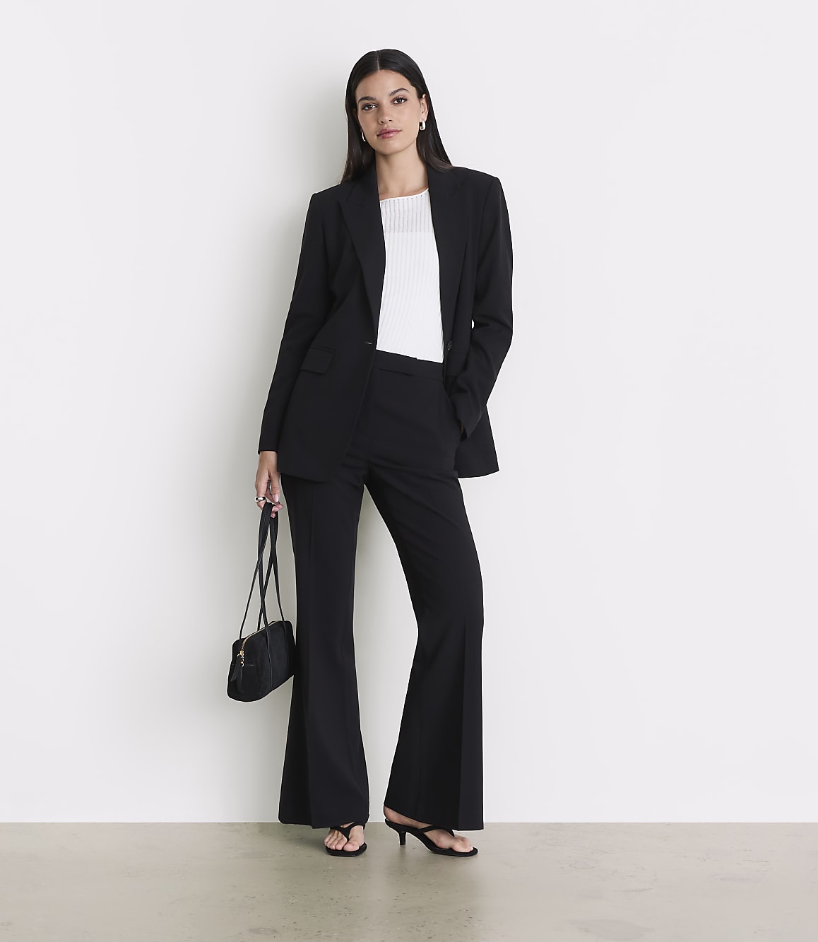 A woman in a black suit and white top stands against a white wall, holding a black purse.
