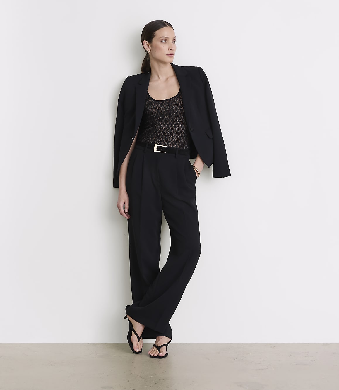 A woman in a black blazer, lace top, and wide-leg pants stands against a plain white wall.