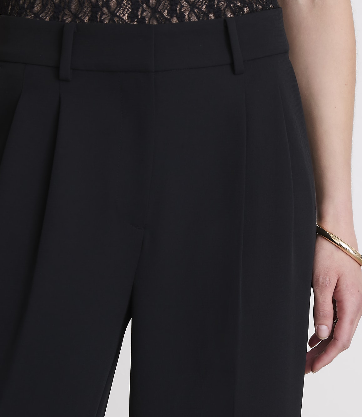 Black pleated trousers are in the foreground, with a person's arm and a gold bracelet visible on the right, and a hint of lace fabric at the top.