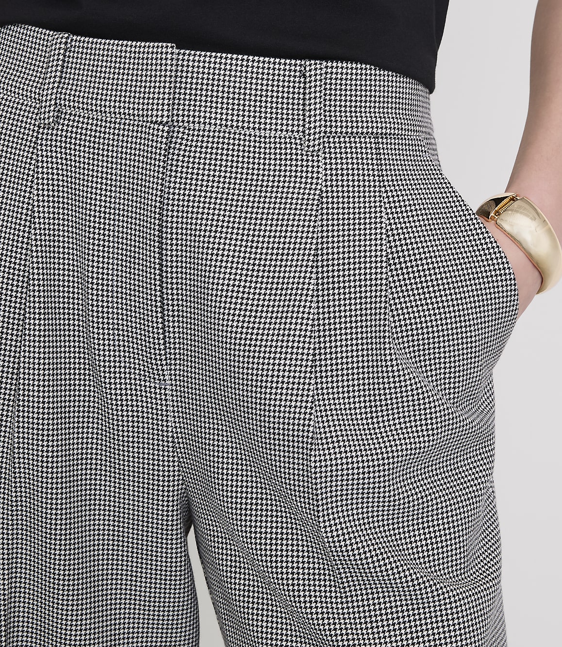 A person's hand, adorned with a gold bracelet, rests in the pocket of houndstooth patterned pants against a plain white background.