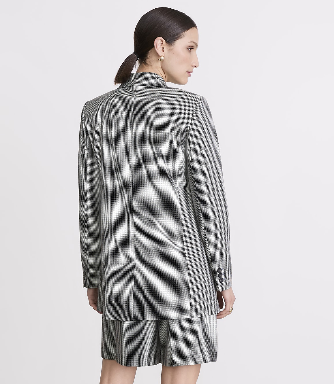 A woman in a checkered blazer and shorts stands against a plain gray background.