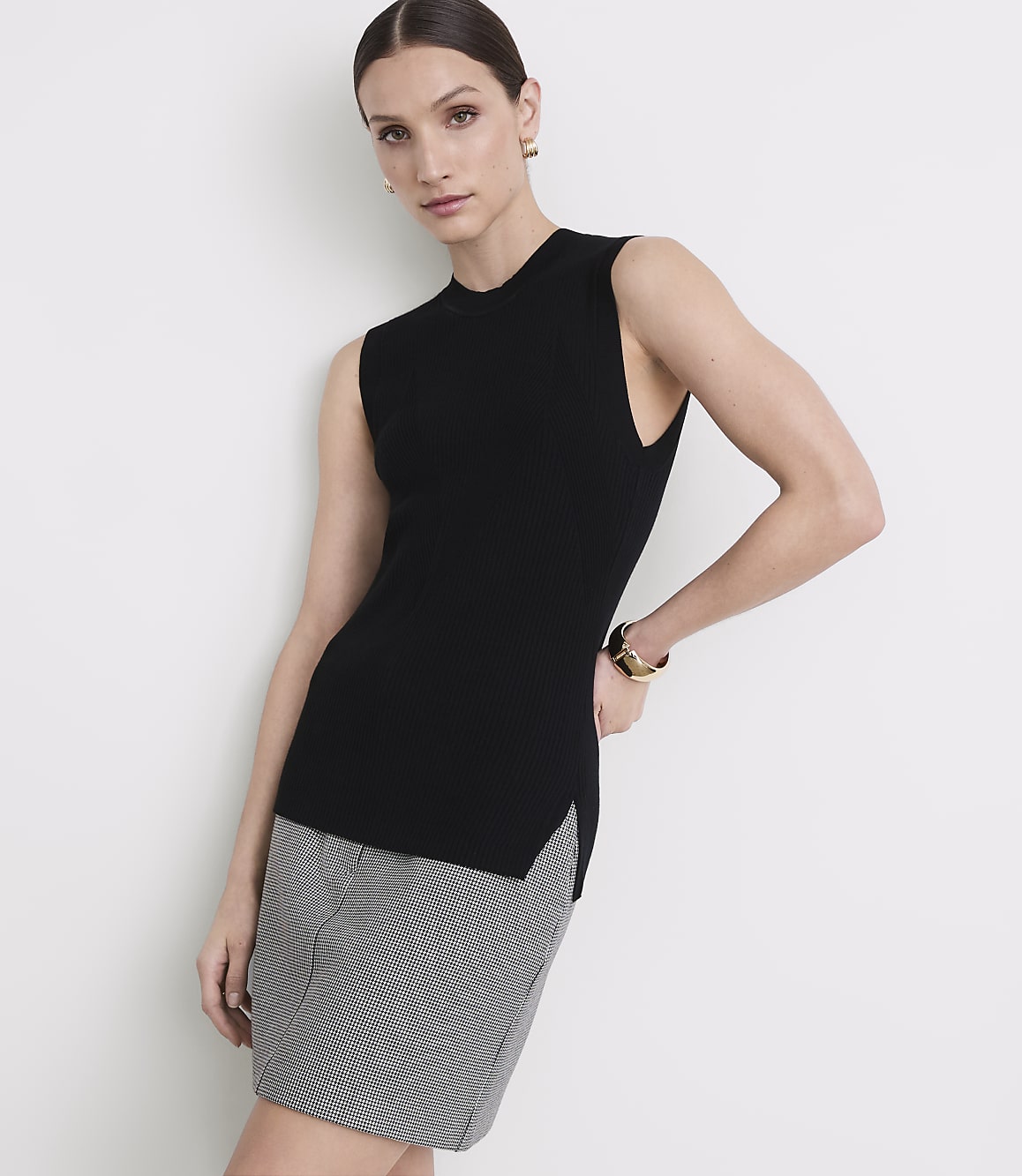 A woman in a black sleeveless top and a patterned skirt stands against a plain white background.