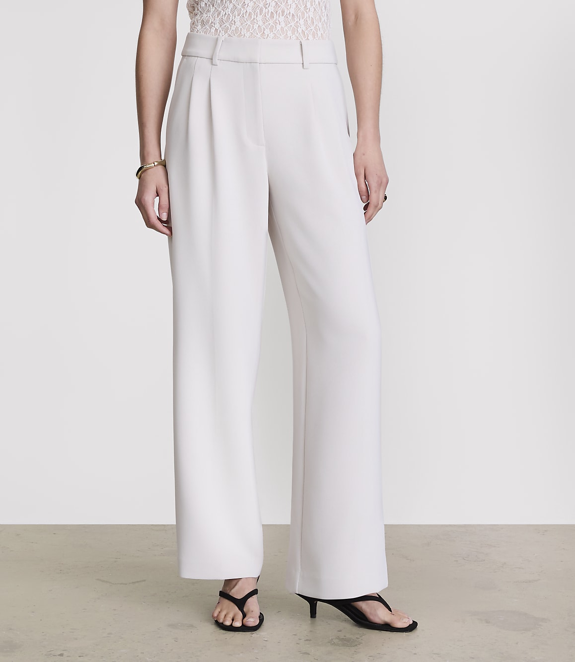 A person stands in white wide-leg trousers and a lace top against a plain grey background.