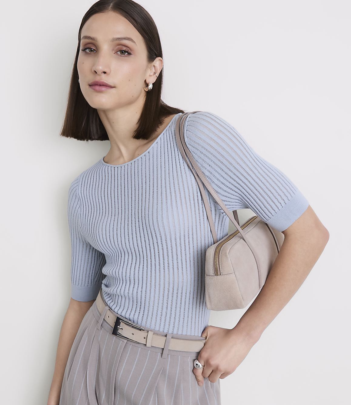 A woman in a light blue ribbed top and pinstripe pants poses with a small beige handbag against a plain white background.