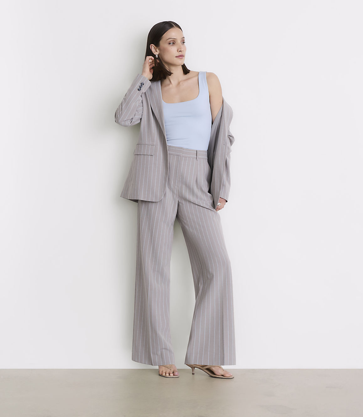 A woman in a light blue tank top and a pinstripe suit stands against a plain white wall.