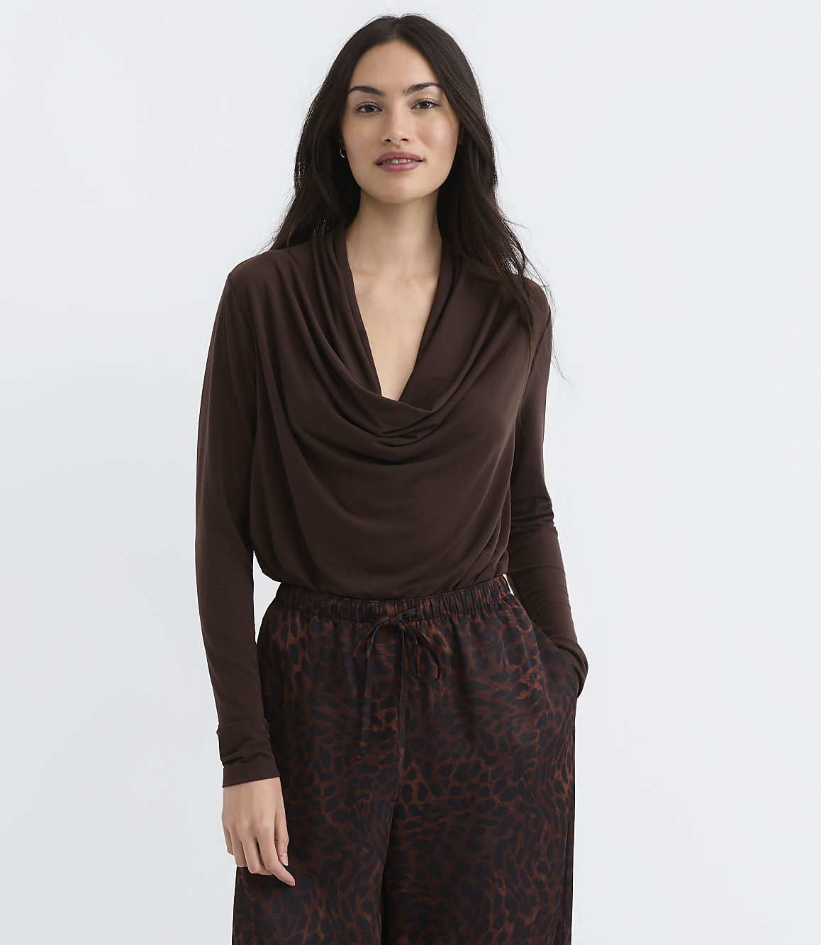 A woman in a brown cowl-neck top and leopard-print pants stands against a plain white background.