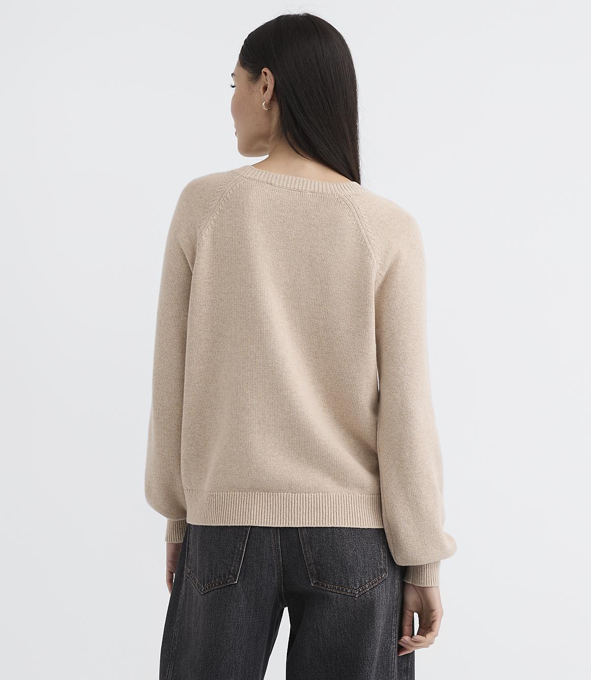 A woman in a beige sweater and dark jeans stands against a plain, light background.