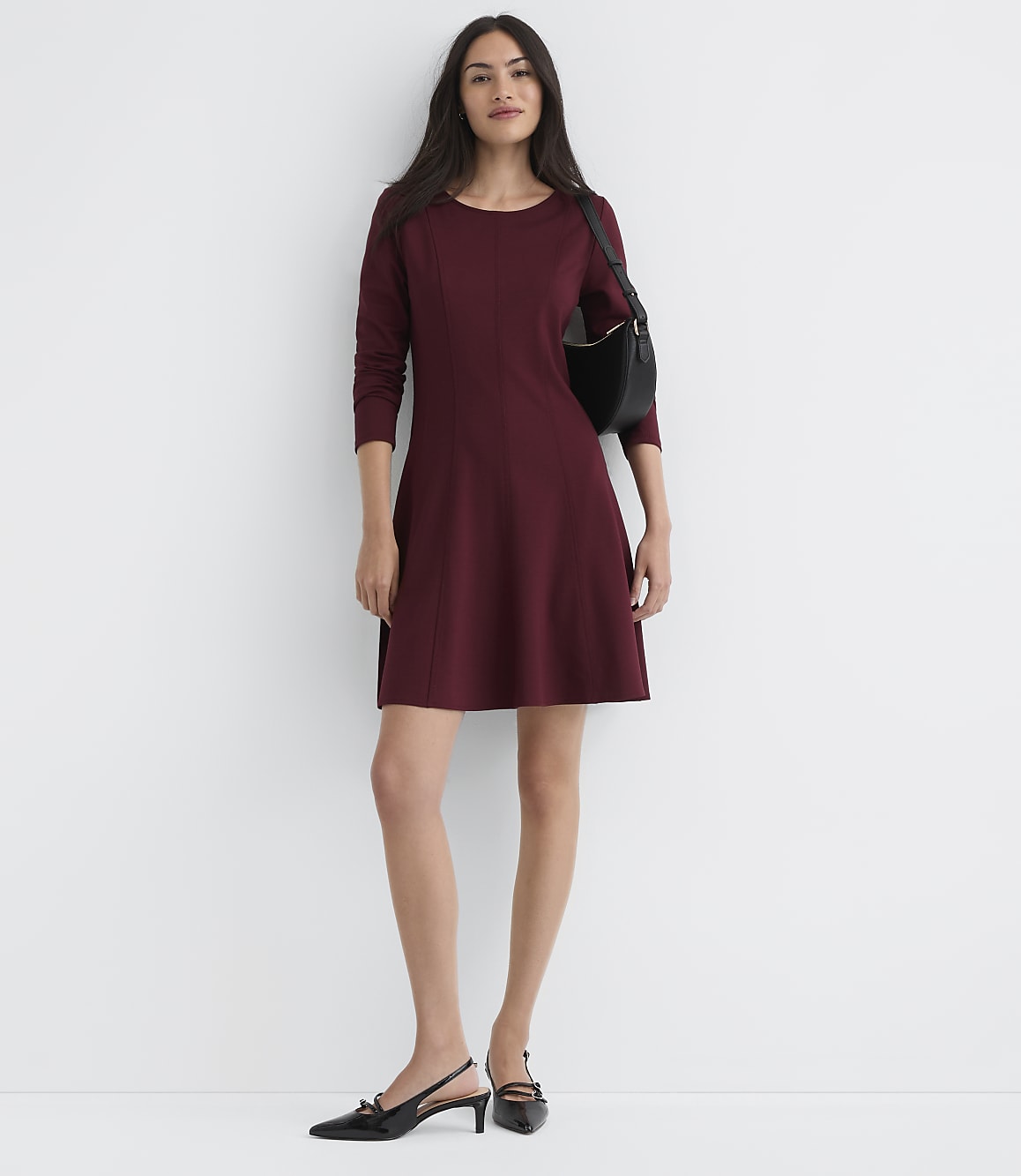 A woman in a maroon dress and black slingback heels stands against a plain white wall, with a black purse slung over her shoulder.