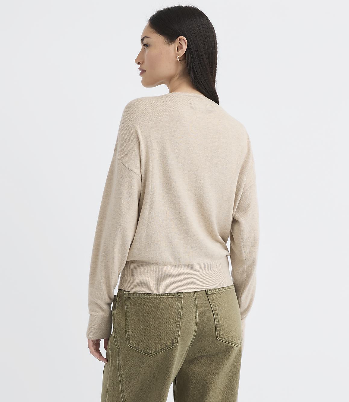 A woman in a beige sweater and olive green pants stands against a plain white background.