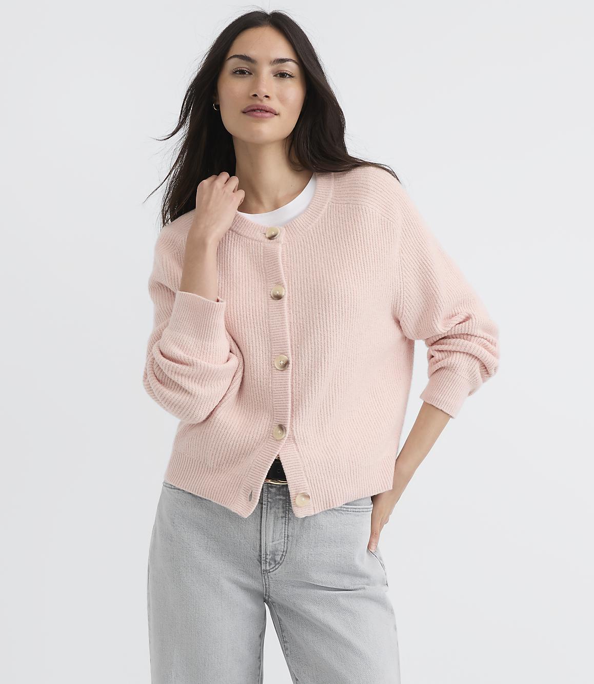 A woman in a light pink cardigan and grey jeans stands against a plain white background.