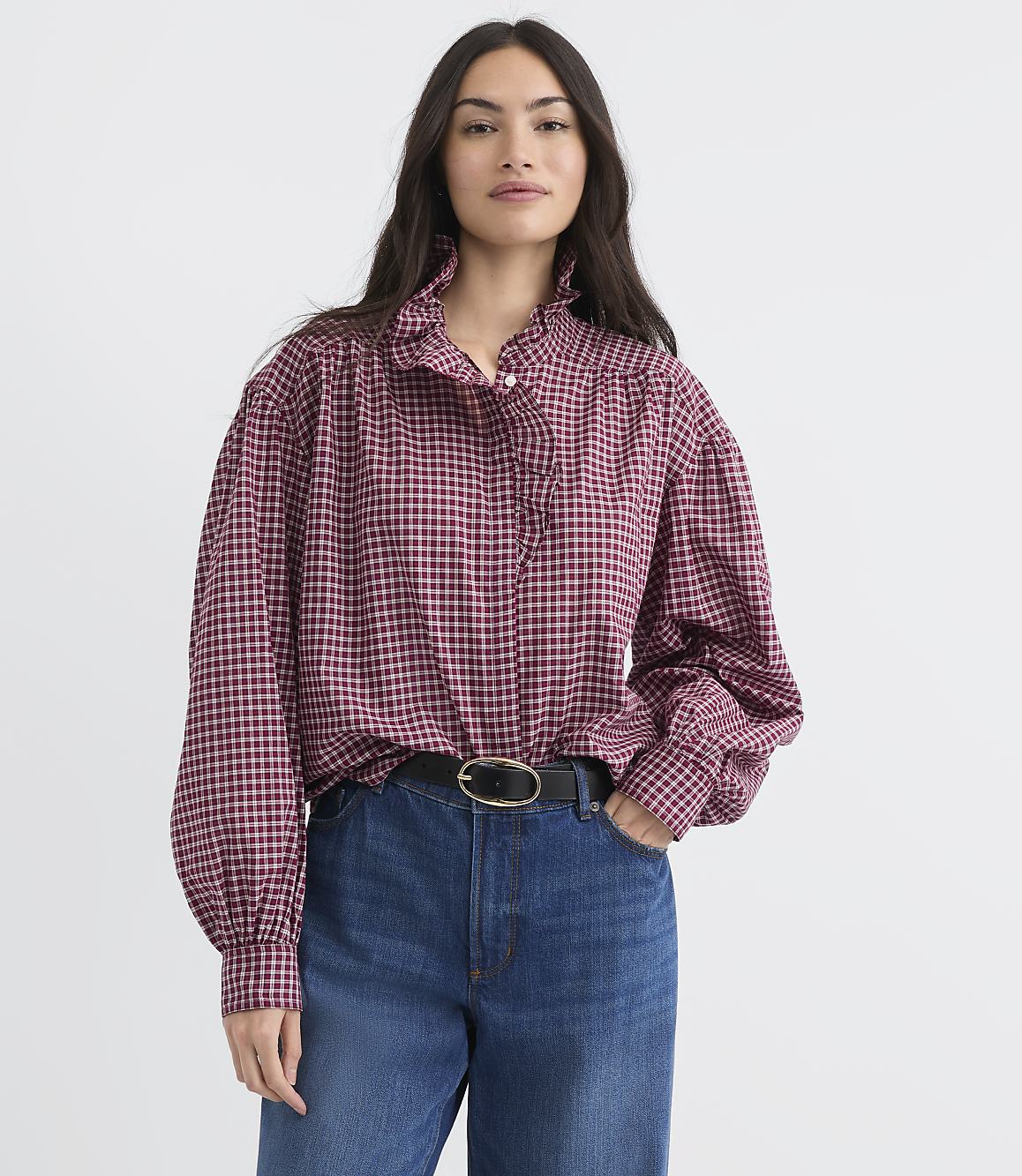 A woman in a ruffled, checkered blouse and jeans stands against a plain white background.