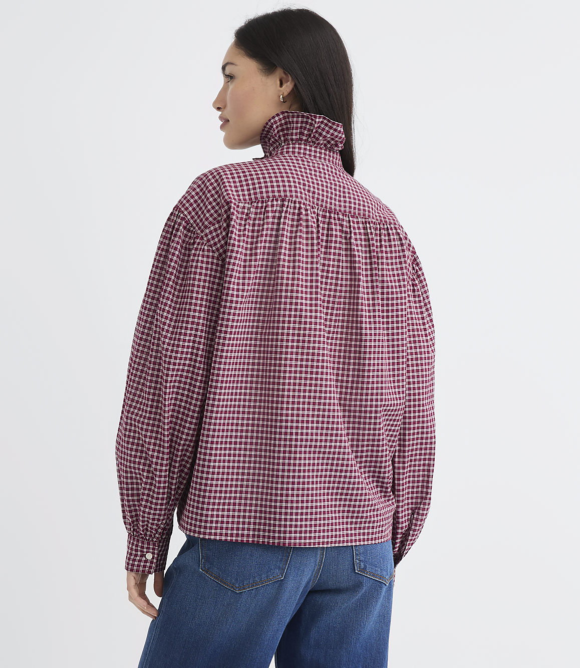 A woman in a red and black checkered blouse with a ruffled collar stands against a plain white background.