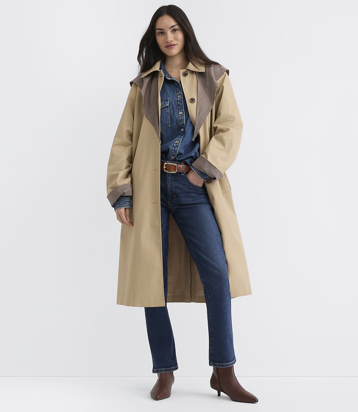 A woman in a tan trench coat, denim shirt, and jeans stands against a plain white background.