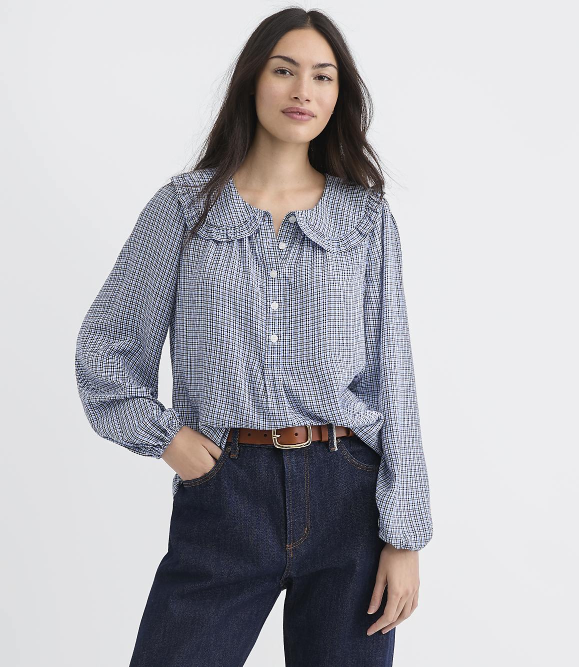 A woman in a blue checkered blouse and dark jeans stands against a plain white background.