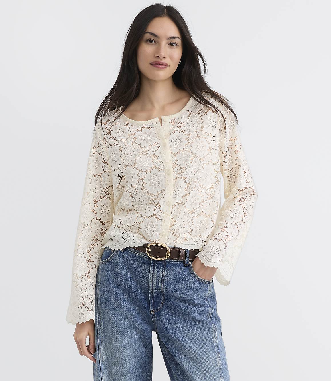 A woman in a cream lace top and blue jeans stands against a plain white background.